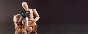 pelear un dwi-Como-Combatir-un-DUI-en-Winston-Salem-Greensboro-High-Point-NC-800×314 Figura de madera sentada con la cabeza inclinada, apoyada en un vaso de licor con un manojo de llaves encima, simbolizando las consecuencias de conducir en estado de ebriedad.